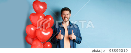 Happy valentines day. Cheerful boyfriend showing thumbs up in approval, standing with red hearts balloons for lover, blue background 98396019
