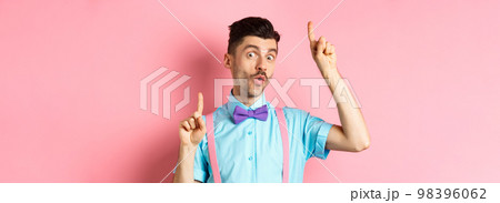 Cheerful funny man dancing in bow-tie and suspenders, pointing fingers up and looking upbeat, standing over pink background Cheerful funny man dancing in bow-tie and suspenders, pointing fingers up and looking upbeat, standing over pink background 98396062