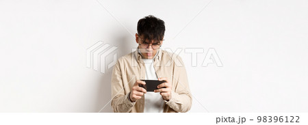 Handsome young man stare at mobile screen and playing video game on smartphone, standing on white background 98396122