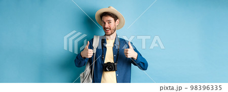 Happy smiling tourist say yes and show thumbs up, going on summer vacation, holding backpack and photo camera, blue background 98396335