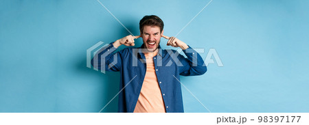 Angry young man shut ears to block annoying sound and shouting at camera, frowning and looking furious, standing on blue background Angry young man shut ears to block annoying sound and shouting at camera, frowning and looking furious, standing on blue background 98397177