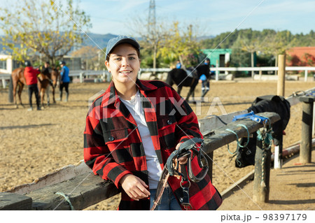 Portrait of positive young female stable worker holding horse halter or harness near wooden fence on ranch Portrait of positive young female stable worker holding horse halter or harness near wooden fence on ranch 98397179