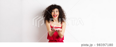 Holidays and celebration concept. Smiling woman in red dress giving you birthday cake with candle, standing over white background Holidays and celebration concept. Smiling woman in red dress giving you birthday cake with candle, standing over white background 98397180
