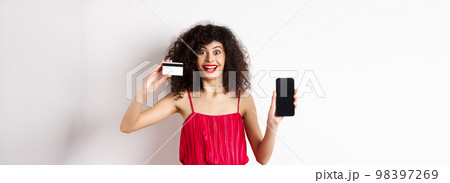 Online shopping concept. Excited curly-haired woman in red dress showing empty smartphone screen and plastic credit card, smiling happy at camera, standing on white background Online shopping concept. Excited curly-haired woman in red dress showing empty smartphone screen and plastic credit card, smiling happy at camera, standing on white background 98397269
