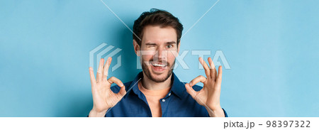 Close up of handsome confident guy winking, showing okay gestures, guarantee quality, recommending something good, standing on blue background Close up of handsome confident guy winking, showing okay gestures, guarantee quality, recommending something good, standing on blue background 98397322