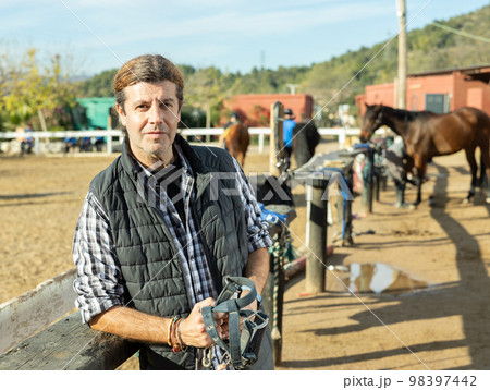 Middle-aged man instructor holding horse halter or reins during working day at horse club on sunny day of spring 98397442