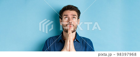 Close up shot of hopeful guy pleading God, holding hands in begging gesture and looking up in sky, standing on blue background supplicating 98397968