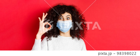 Health, pandemic and covid concept. Excited woman show yes okay sign, wearing medical mask and looking with approval, standing against red background Health, pandemic and covid concept. Excited woman show yes okay sign, wearing medical mask and looking with approval, standing against red background 98398022