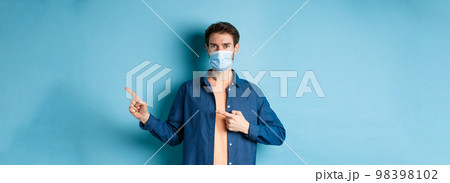 Covid and healthcare concept. Handsome modern guy in medical mask pointing at empty space aside, standing on blue background Covid and healthcare concept. Handsome modern guy in medical mask pointing at empty space aside, standing on blue background 98398102