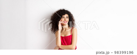 Elegant and romantic woman in red dress and makeup, smiling and touching her face, looking aside with happy expression, white background 98399196