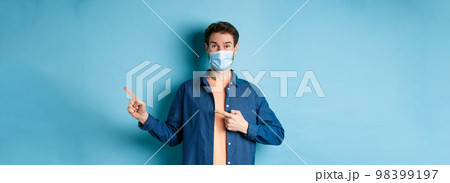Covid-19 and pandemic concept. Happy healthy guy in medical mask pointing fingers at upper right corner, showing advertisement, standing on blue background 98399197