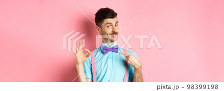 Image of young handsome guy in bow-tie and holiday outfit, pointing at himself and showing okay gesture, self-promoting on pink background 98399198
