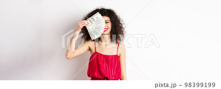 Excited woman in red dress winning money, showing dollar bills and smiling happy, standing on white background 98399199