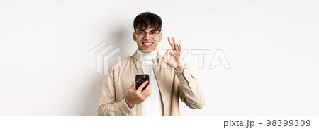 Technology and online shopping concept. Portrait of handsome modern guy in glasses showing OK gesture using smartphone, recommending app or shop, white background 98399309