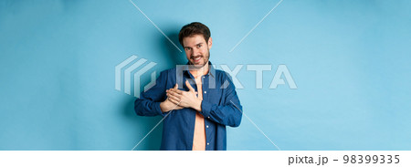 Smiling man feeling touched and grateful, holding hands on heart and thanking, standing on blue background 98399335