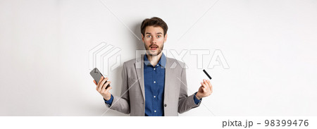 Online shopping. Confused man in suit spread hands sideways, holding plastic credit card with mobile phone and shrugging cueless, standing on white background Online shopping. Confused man in suit spread hands sideways, holding plastic credit card with mobile phone and shrugging cueless, standing on white background 98399476