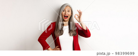 Happy asian mature woman in red blazer showing okay sign, smiling amazed, praising something awesome, standing over white background Happy asian mature woman in red blazer showing okay sign, smiling amazed, praising something awesome, standing over white background 98399699