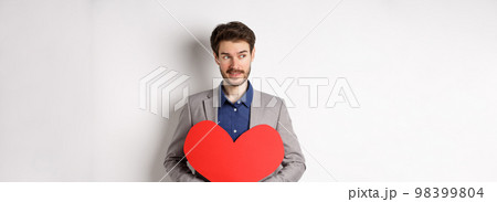 Attractive man in suit looking left and smiling, holding red heart cutout, prepare valentines day surprise for lover, standing over white background 98399804