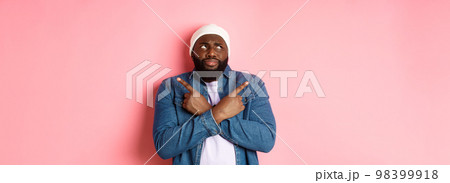 Confused Black man with beard, making choice, pointing fingers sideways and looking puzzled, standing over pink background 98399918