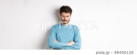 Image of offended and sad young man with beard, look from under forehead and sulking irritated, cross arms on chest defensive, mad at someone, standing on white background 98400126