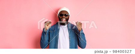 Satisfied african-american man in sunglasses saying yes, triumphing and making fist pumps in rejoice, celebrating achievement, standing like winner over pink background 98400327