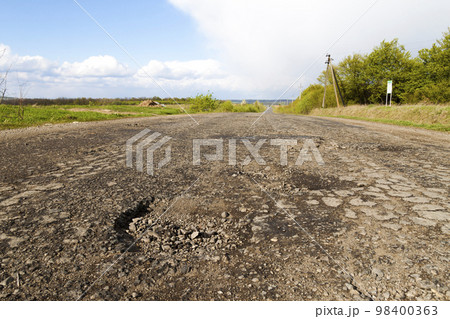 Damaged rural road, cracked asphalt blacktop with potholes and patches. Poor quality of road repair work. Damaged rural road, cracked asphalt blacktop with potholes and patches. Poor quality of road repair work. 98400363