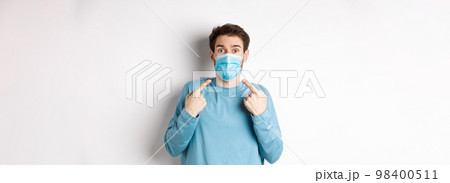 Coronavirus, health and quarantine concept. Confused guy pointing at his medical mask and looking surprised at camera, standing over white background 98400511