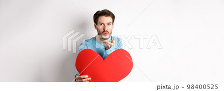 Romantic boyfriend sending air kiss at camera and holding big red Valentines day heart, standing over white background 98400525
