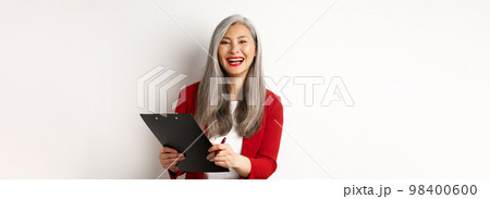 Happy asian office employee, woman in red blazer, working and laughing, holding clipboard with documents, standing over white background Happy asian office employee, woman in red blazer, working and laughing, holding clipboard with documents, standing over white background 98400600