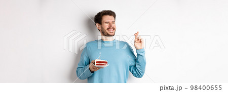Celebration and holidays concept. Young man celebrating birthday, cross fingers for good luck, making wish on bday cake with lit candle, standing over white background 98400655