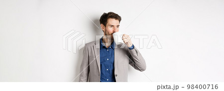 Handsome businessman in suit drinking coffee or tea from office mug, looking aside at logo, standing against white background Handsome businessman in suit drinking coffee or tea from office mug, looking aside at logo, standing against white background 98400716