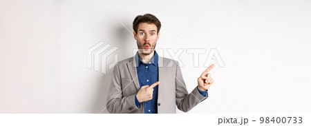 Excited handsome man in suit pointing fingers left at banner and saying wow, look here gesture, showing advertisement, standing on white background 98400733
