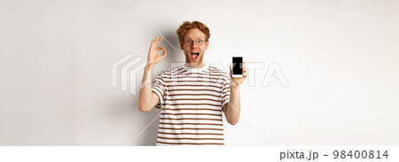 Technology and e-commerce concept. Young man with red hair showing okay sign and blank smartphone screen, praising awesome app, standing over white background 98400814