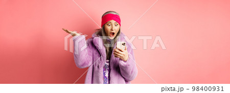 Online shopping and fashion concept. Amazed and surprised asian senior woman looking at smartphone screen, reading on mobile phone and gasping, pink background 98400931