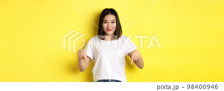 Beauty and fashion concept. Beautiful asian woman in white t-shirt pointing fingers down, demonstrate logo standing over yellow background Beauty and fashion concept. Beautiful asian woman in white t-shirt pointing fingers down, demonstrate logo standing over yellow background 98400946