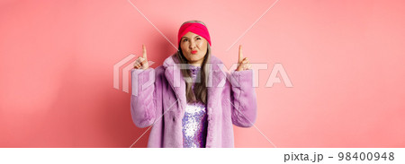 Stylish middle aged asian woman in purple fake fur coat, looking skeptical and grimacing with displeased face, pointing fingers up at something bad and unimpressive, pink background 98400948