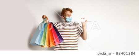 Pandemic and special offer concept. Happy redhead man in face mask showing shopping bags and plastic credit card, standing over white background Pandemic and special offer concept. Happy redhead man in face mask showing shopping bags and plastic credit card, standing over white background 98401384