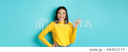 Impressed asian woman in yellow sweater, looking left amazed and showing OK sign in approval, standing over blue background Impressed asian woman in yellow sweater, looking left amazed and showing OK sign in approval, standing over blue background 98401574