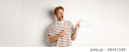 Check it out. Amazed redhead guy pointing and looking left at promotion banner, standing over white background 98401586