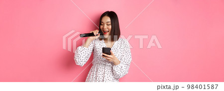 Cute asian woman reading lyrics in smartphone, singing with microphone, standing in trendy dress over pink background Cute asian woman reading lyrics in smartphone, singing with microphone, standing in trendy dress over pink background 98401587