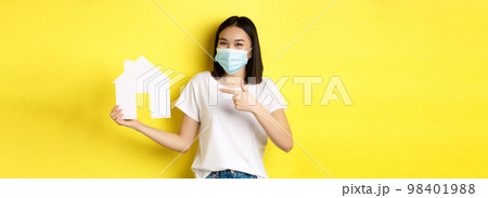 Covid-19, pandemic and real estate concept. Cheerful asian woman smiling in medical mask, showing paper house cutout, recommend agency for buying property 98401988