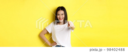 We need you. Confident asian woman smiling, pointing finger at camera, inviting to join her, beckon or choosing someone, standing over yellow background 98402488