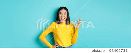 Impressed asian woman in yellow sweater, looking left amazed and showing OK sign in approval, standing over blue background Impressed asian woman in yellow sweater, looking left amazed and showing OK sign in approval, standing over blue background 98402731