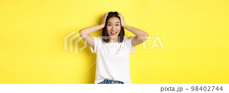Amazed asian girl winning prize and looking with excitement and disbelief, holding hands on head and smiling happy, standing over yellow background 98402744