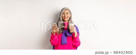 E-commerce concept. Beautiful asian senior woman drinking coffee from takeaway cup and using mobile phone, smiling at camera, white background 98402900