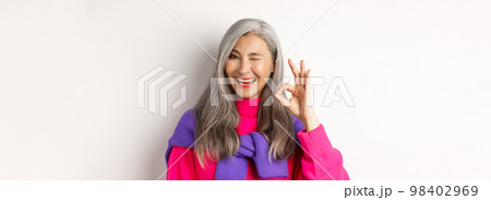 Close-up of cheerful asian grandmother in fashionable pink sweater, winking and showing okay sign, recommending promotion, standing over white background 98402969