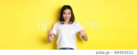Beauty and fashion concept. Beautiful asian woman in white t-shirt pointing fingers down, demonstrate logo standing over yellow background 98403012