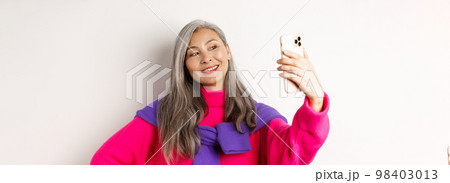 Beautiful and stylish asian senior woman taking selfie on smartphone, smiling and posing in trendy pink sweater, standing over white background Beautiful and stylish asian senior woman taking selfie on smartphone, smiling and posing in trendy pink sweater, standing over white background 98403013