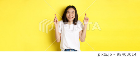 Hopeful asian girl smiling optimistic, feeling lucky, cross fingers and making wish, standing over yellow background Hopeful asian girl smiling optimistic, feeling lucky, cross fingers and making wish, standing over yellow background 98403014