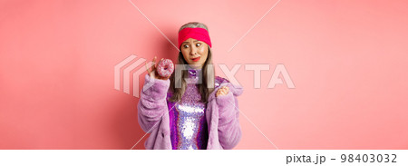 Funny asian senior woman in glittering disco dress and faux fur coat looking tempted at delicious donut, wanting to eat sweet, standing over pink background Funny asian senior woman in glittering disco dress and faux fur coat looking tempted at delicious donut, wanting to eat sweet, standing over pink background 98403032